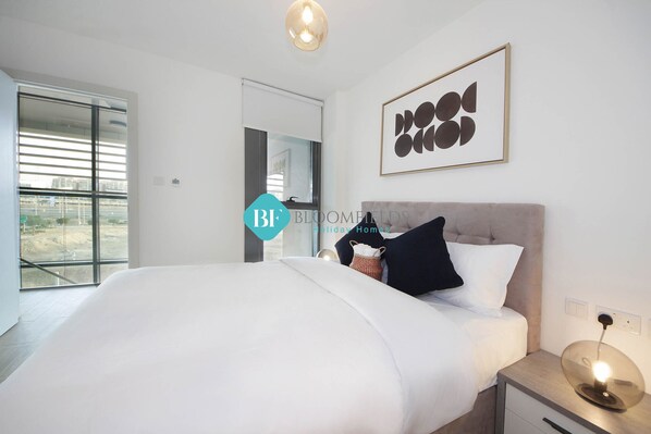 A modern bedroom with a large bed, a bedside table with a lamp, a framed artwork on the wall, and a view of the outdoors through a sliding door.