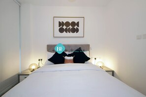 A neatly made bed with white bedding, two bedside lamps, a framed artwork above the bed, and a small basket on the nightstand.