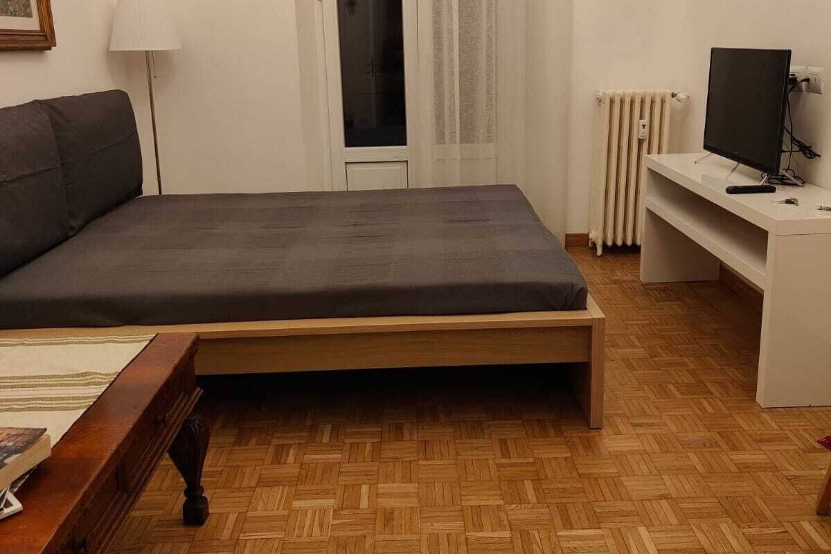 A bedroom with a bed, a desk, a chair, a lamp, a radiator, and a television.