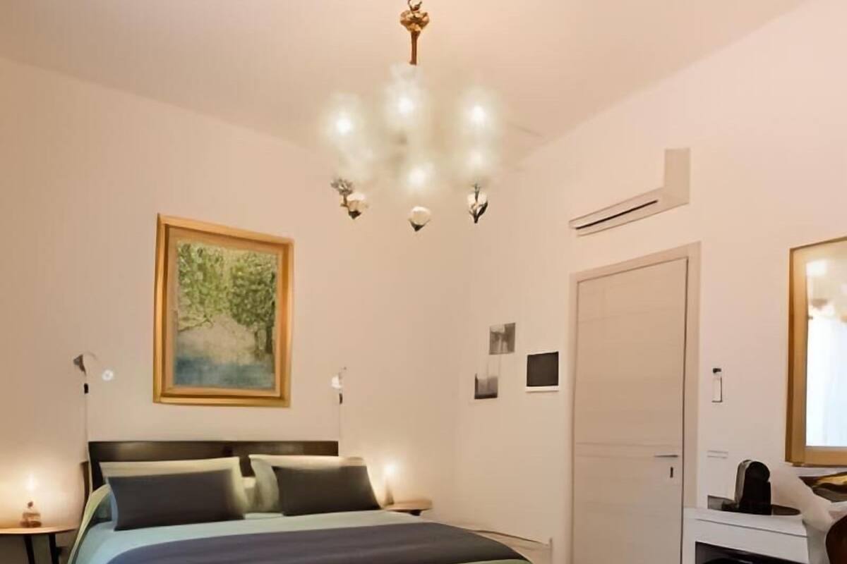 A bedroom with a bed, bedside tables, a desk, a chair, a painting, and a chandelier.