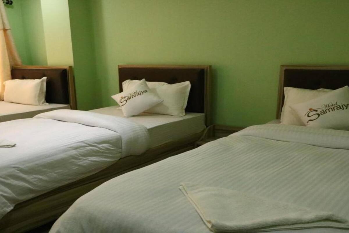 A hotel room with three single beds, each with a pillow labeled "Samrajya." The room has green walls and a window with curtains.