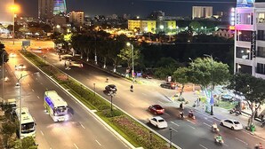 A nighttime cityscape with illuminated buildings, a busy road with vehicles, and a pedestrian area with trees and street lamps. - Rivera Suites (Da Nang)