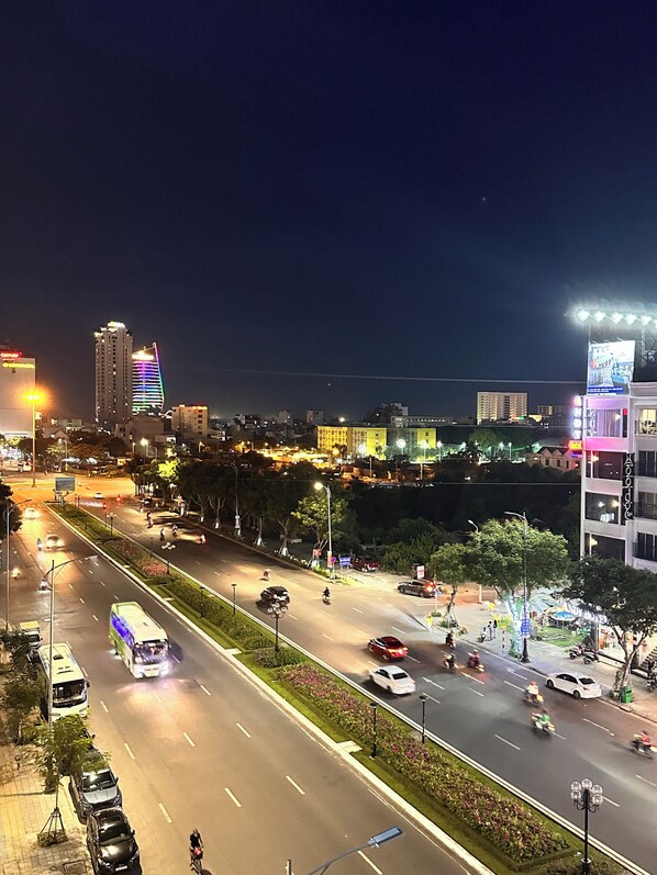 A nighttime cityscape with illuminated buildings, a busy road with vehicles, and a pedestrian area with trees and street lamps. - Rivera Suites (Da Nang)