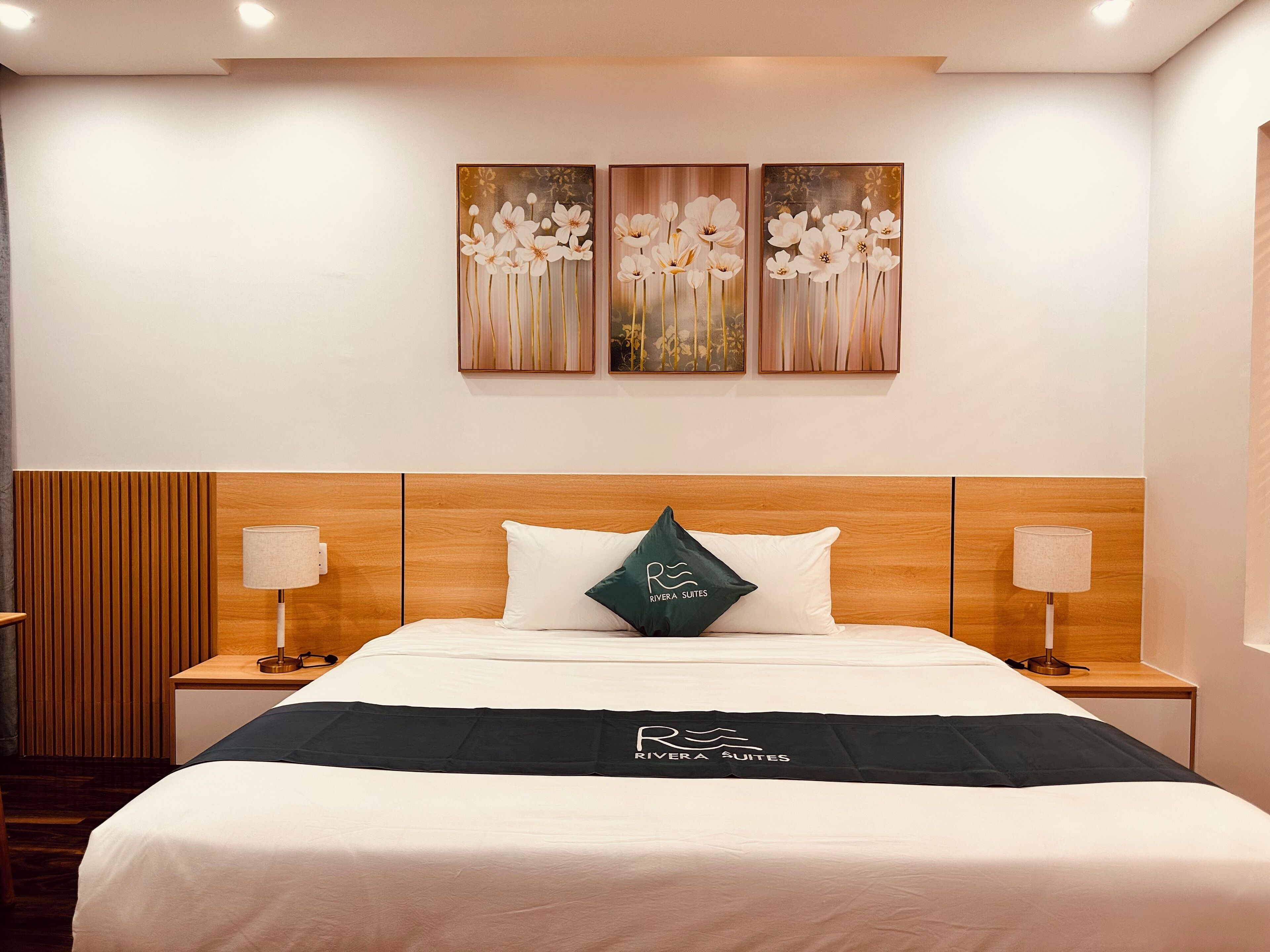 A hotel room with a wooden headboard, a bed with white bedding, two bedside lamps, and three wall-mounted floral paintings.