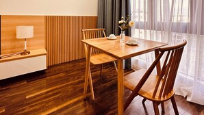 A modern dining area with a wooden table and chairs, a small side table with a lamp, and a view through sheer curtains. - Rivera Suites (Da Nang)