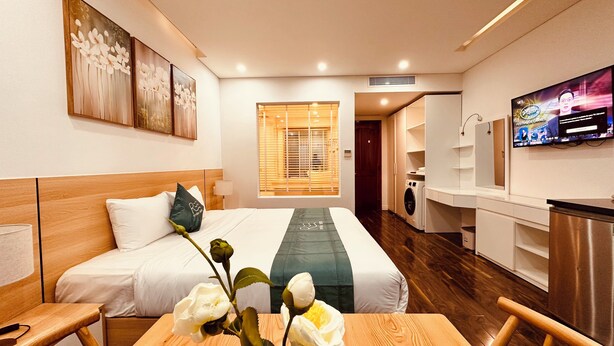 A hotel room with a bed, a wooden table with a vase of flowers, a television, a washing machine, and a mirror. - Rivera Suites (Da Nang)