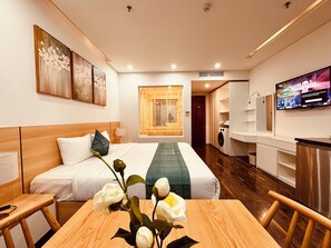 A hotel room with a bed, a wooden table with a vase of flowers, a television, a washing machine, and a mirror. - Rivera Suites (Da Nang)