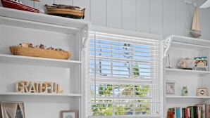 Quarto com paredes de madeira branca, uma janela com persianas e estantes com objetos decorativos, incluindo um modelo de veleiro, conchas e livros.