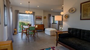 A living room with a dark sofa, a dining area with chairs and a table, a white ottoman, and a view of the outdoors. - Laje da Bica Agroturismo (Covilha)