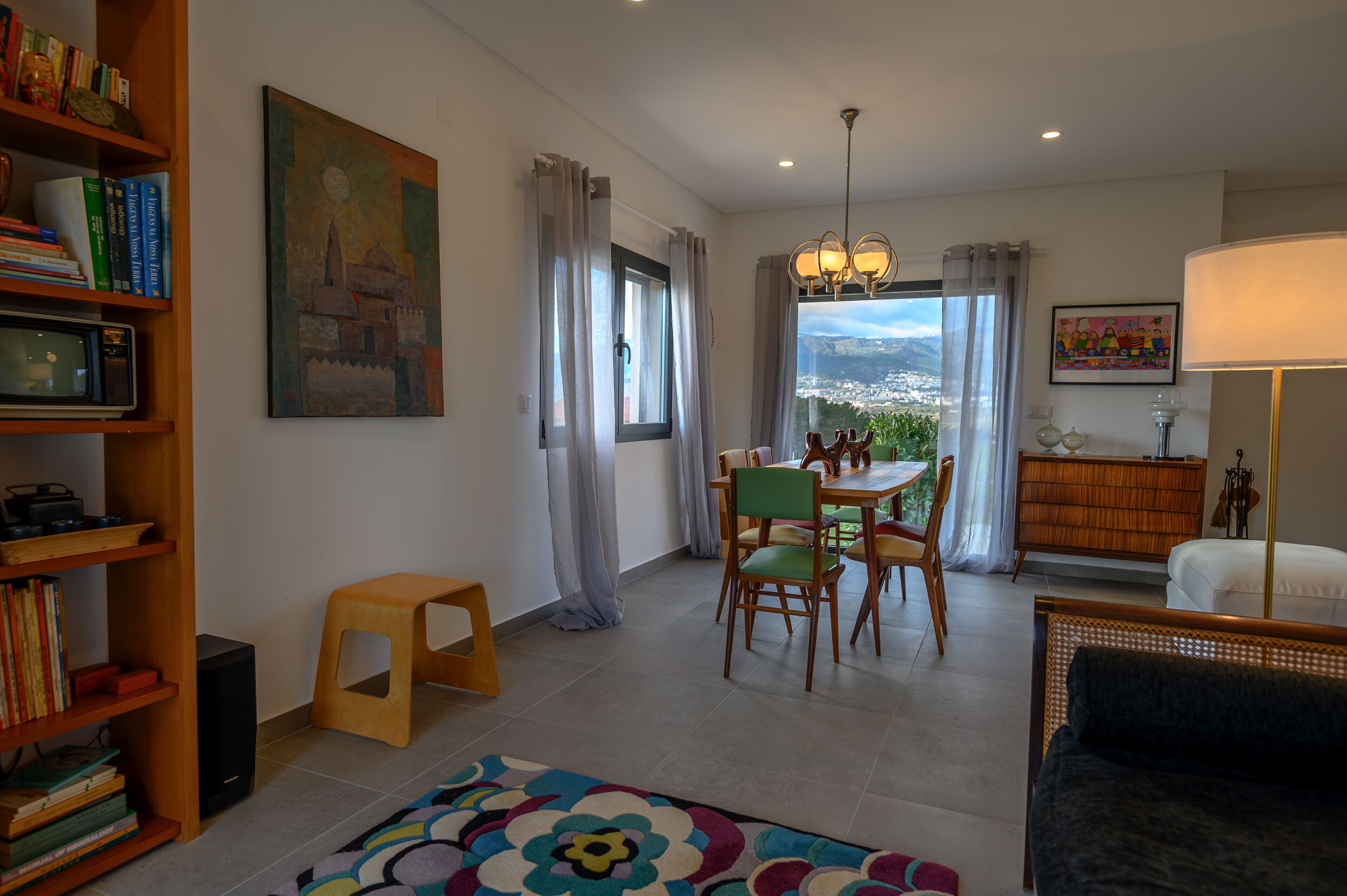 A living room with a dining area, a bookshelf, a painting, and a view of the city.