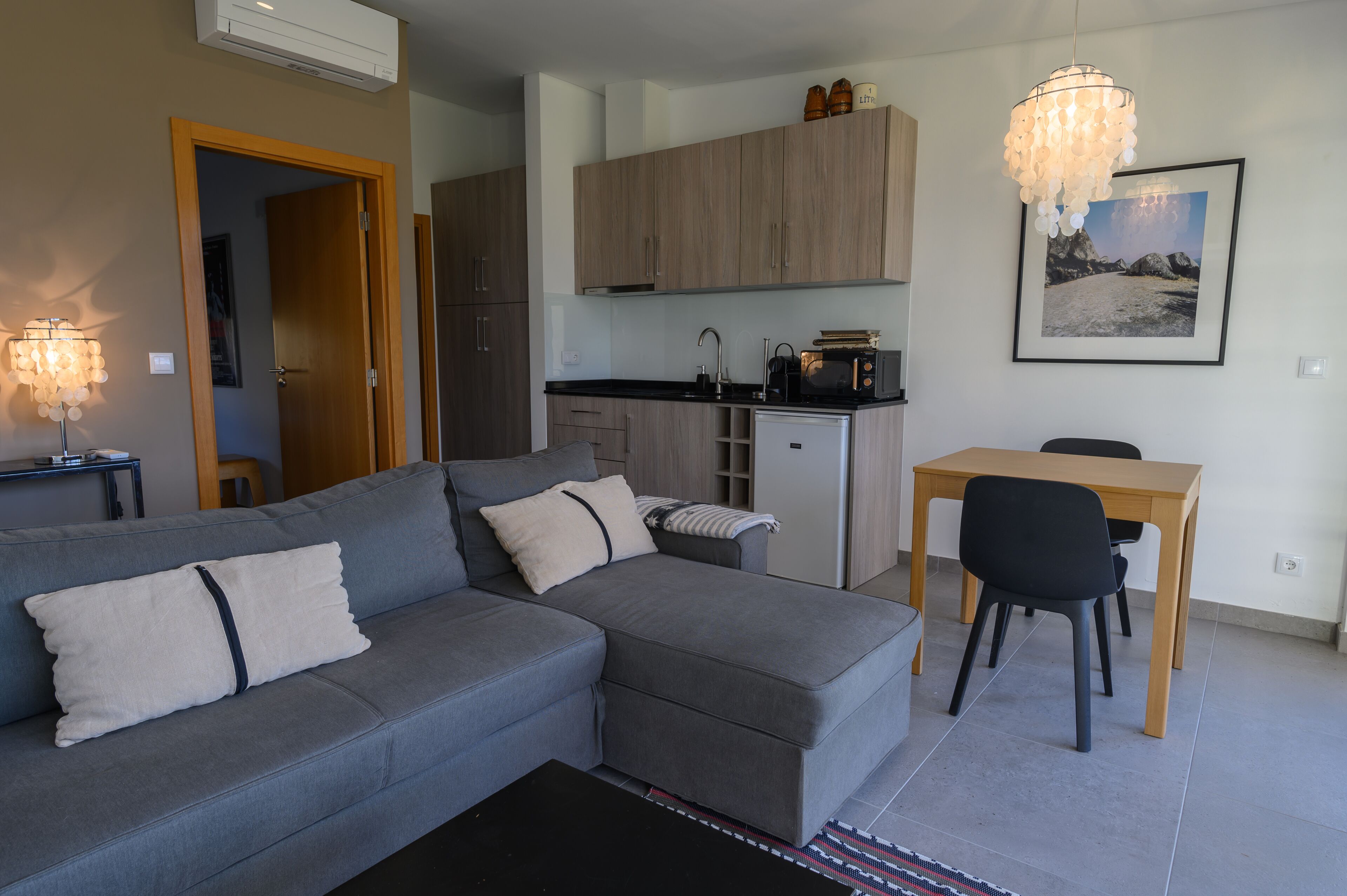 A modern living room with a grey sectional sofa, a dining table with two chairs, a kitchen area with wooden cabinets, and a pendant light.
