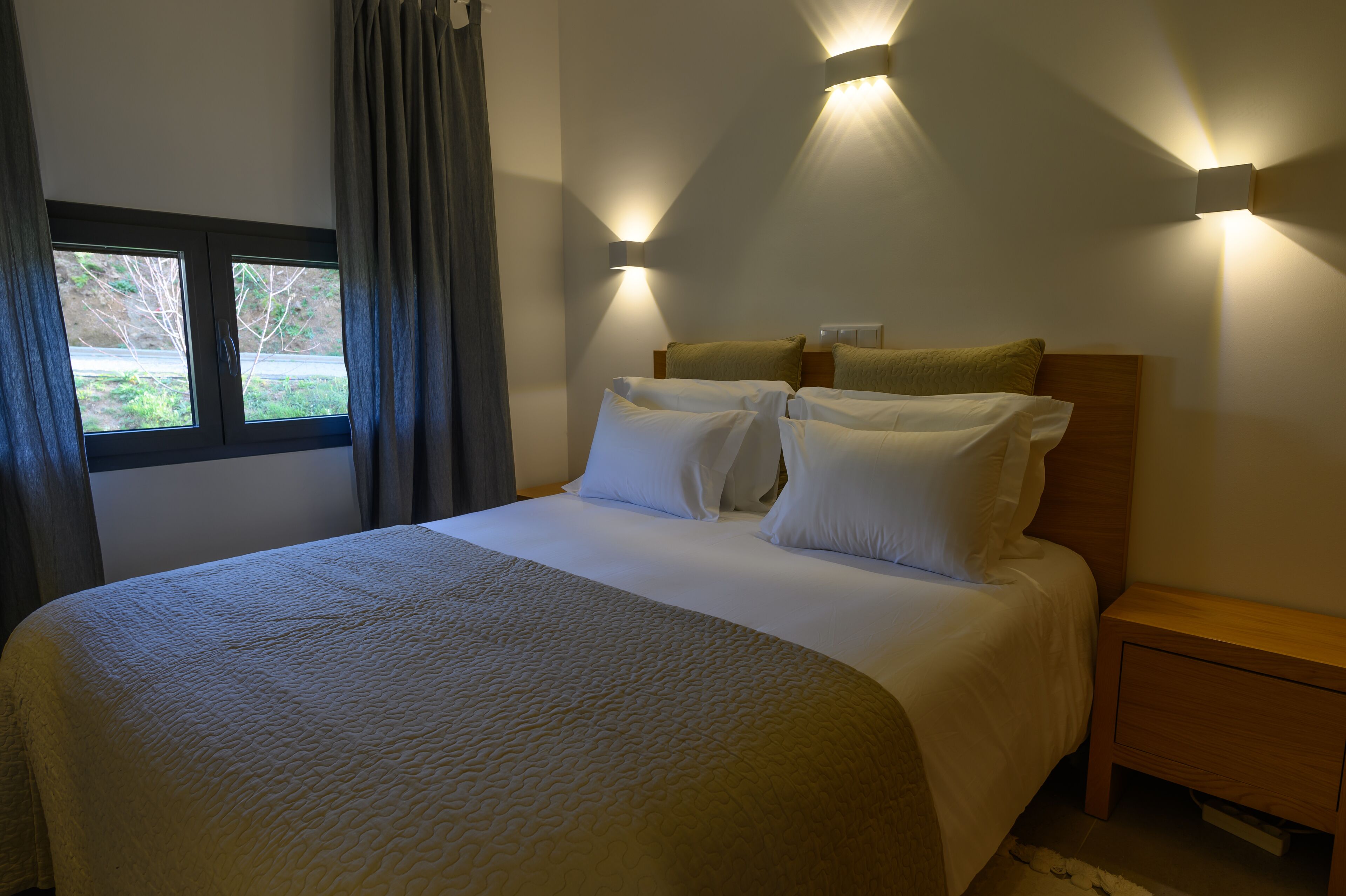 A neatly made bed with white linens and a beige quilt, a wooden nightstand with a drawer, and two wall-mounted light fixtures.