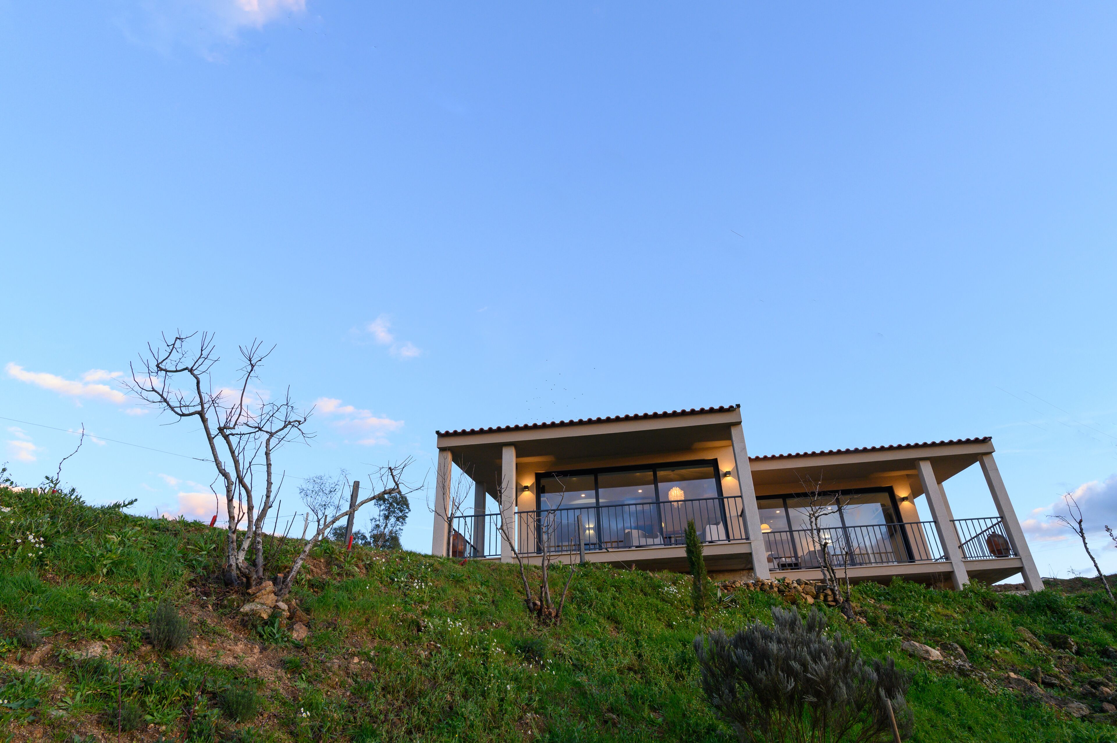 A modern two-story house with large windows and a balcony, situated on a grassy hill with sparse vegetation.