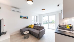 Un salon moderne avec un canapé gris, une petite table d’appoint ronde et un coin cuisine équipé d’une cuisinière et d’ustensiles.