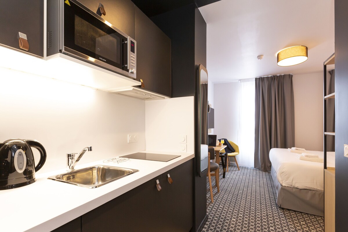 A compact hotel room with a kitchenette, a bed, a small table, and a chair.