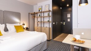 A hotel room with a bed, bedside table, lamp, and a desk with a laptop and croissant. - Appart Hotel Odalys City Le Havre Les Docks (Le Havre)