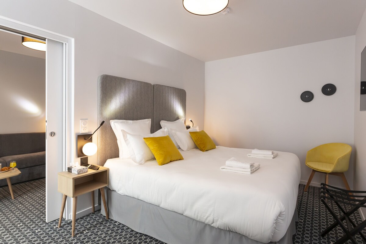 A modern hotel room with a large bed, bedside tables, a chair, and a sofa.