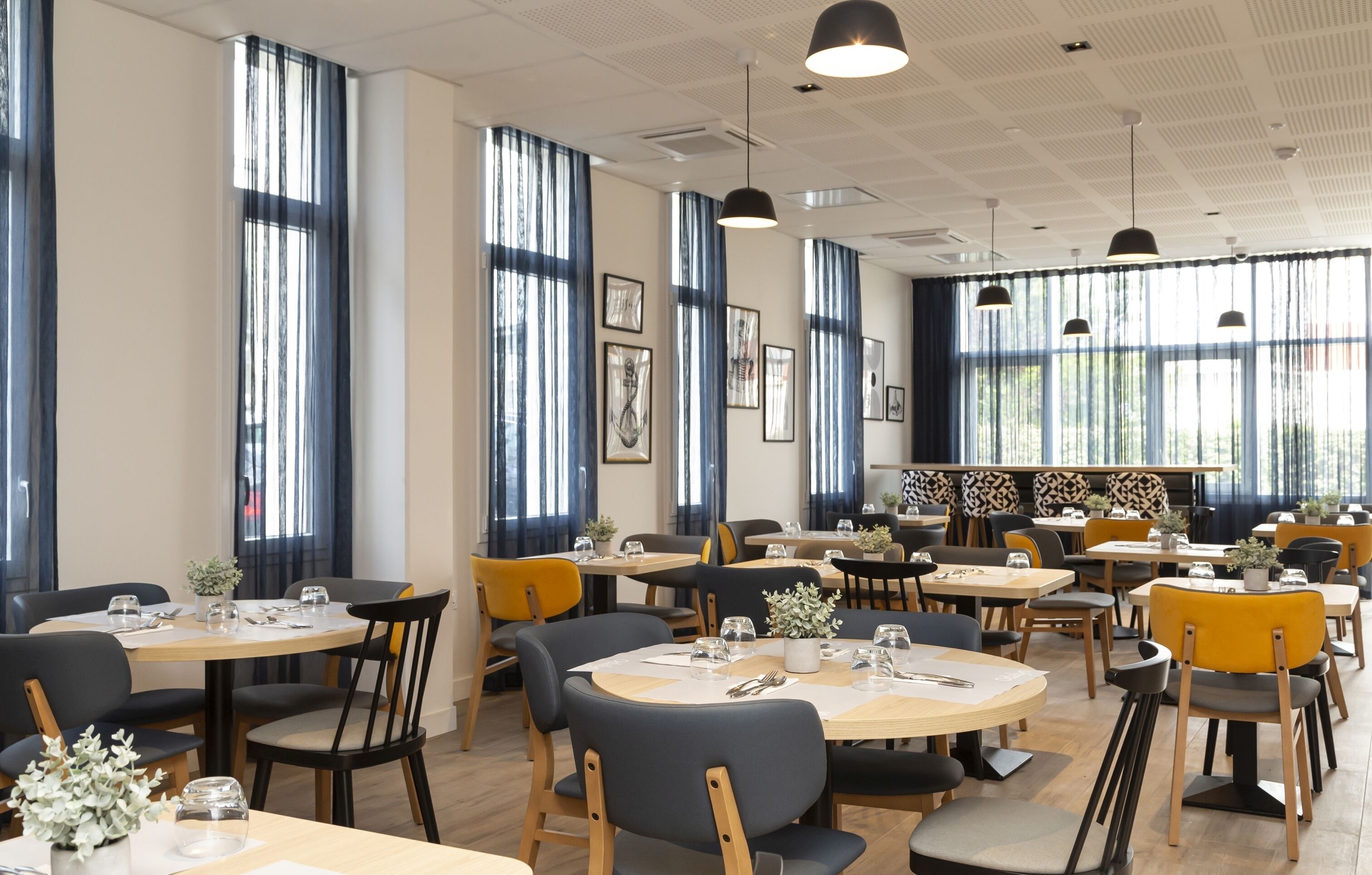 Un comedor amplio con mesas y sillas redondas, ventanas grandes con cortinas e iluminación colgante.