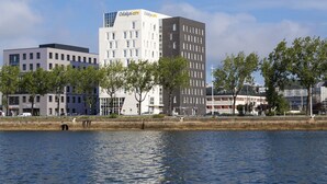 A modern hotel with a prominent 'Odalis City' sign, situated by a waterfront with trees and other buildings in the background. - Appart Hotel Odalys City Le Havre Les Docks (Le Havre)