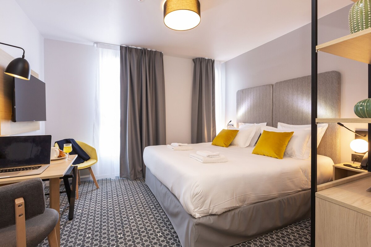 Une chambre d’hôtel avec un lit, un bureau avec un ordinateur portable, une chaise, une table de chevet avec une lampe et une fenêtre avec des rideaux.