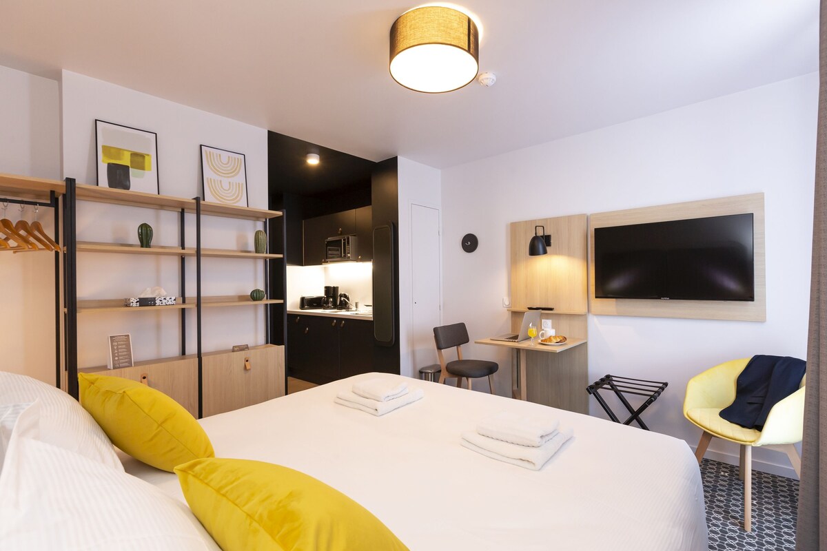 A hotel room with a bed, a desk, a chair, a TV, and a shelf with decorative items.