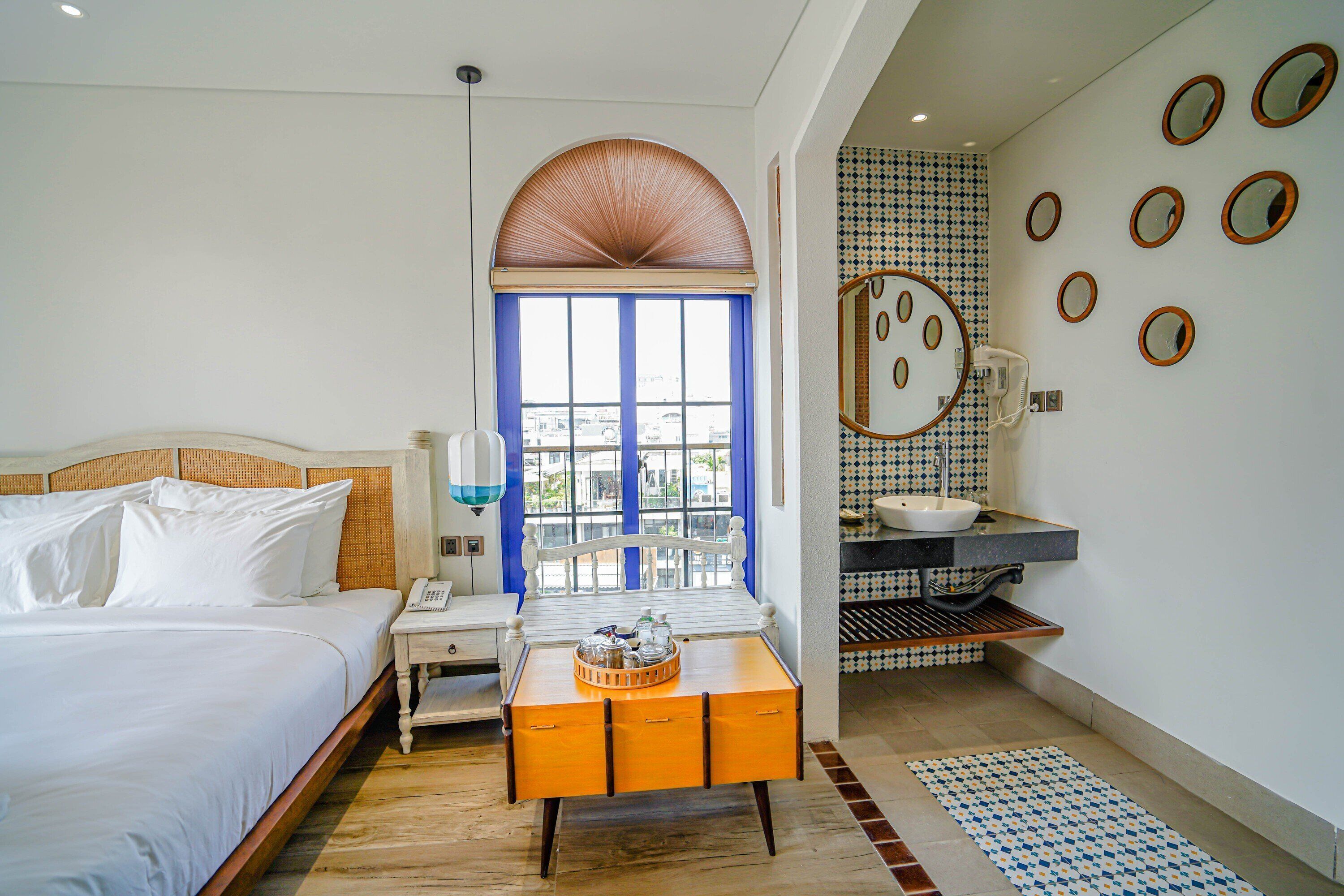 A hotel room with a bed, a small table with a tray, a window with blue curtains, a bathroom with a sink and mirror, and a patterned rug.