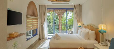 A hotel room with a large bed, a TV, a ceiling fan, a window with curtains, and a view of greenery outside.