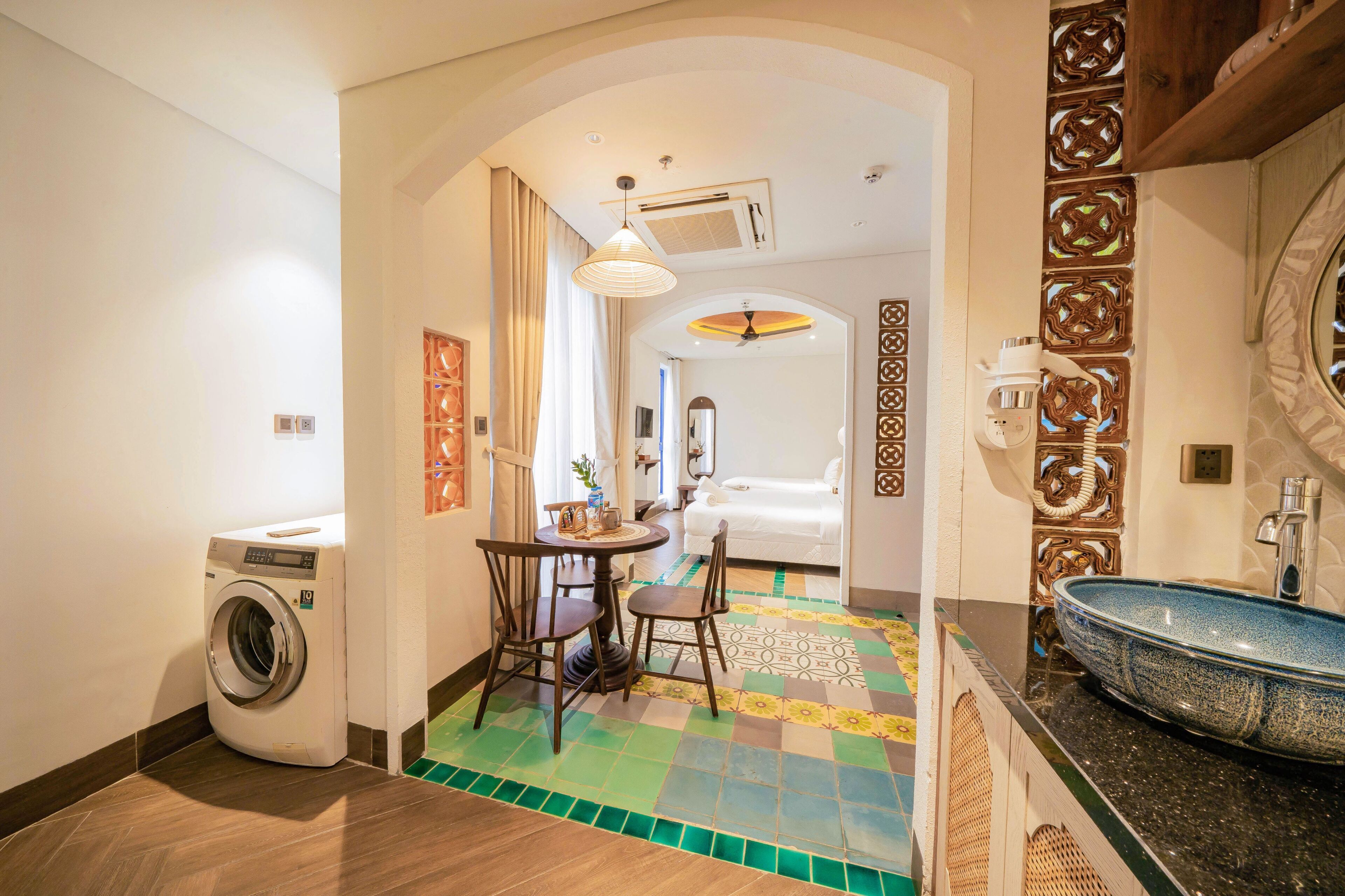 A hotel room with a washing machine, a dining area with chairs and a table, a bathroom with a sink and mirror, and a decorative wall with intricate patterns.
