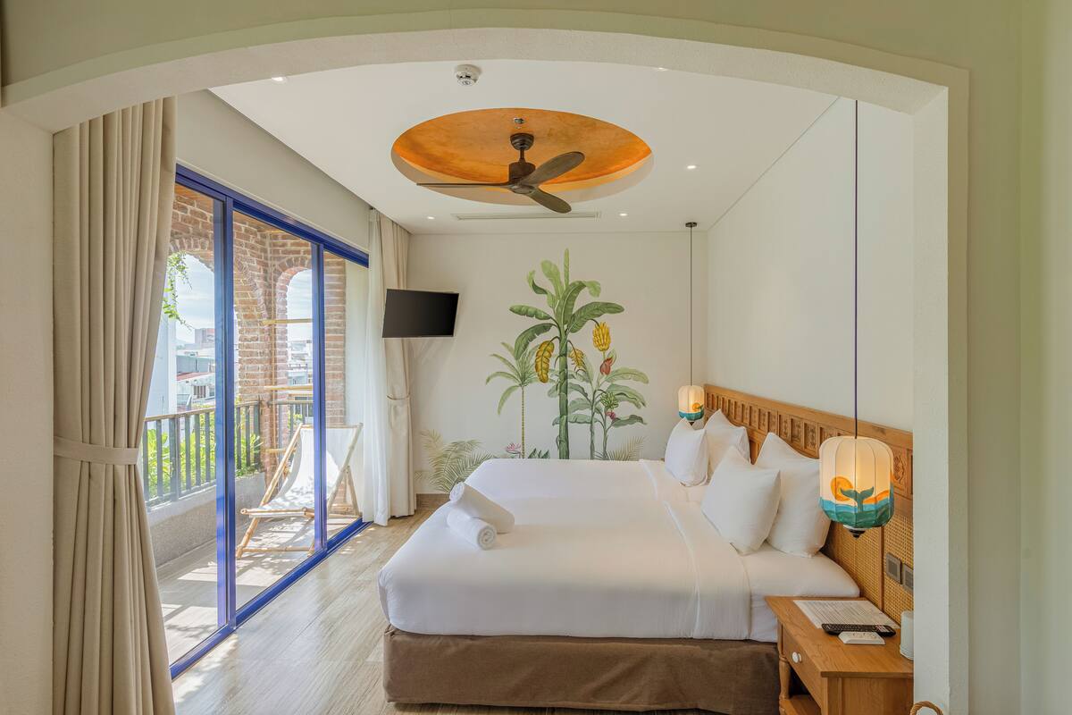 Habitación de hotel con una cama grande, ventilador de techo, televisión y balcón con vistas a la ciudad.