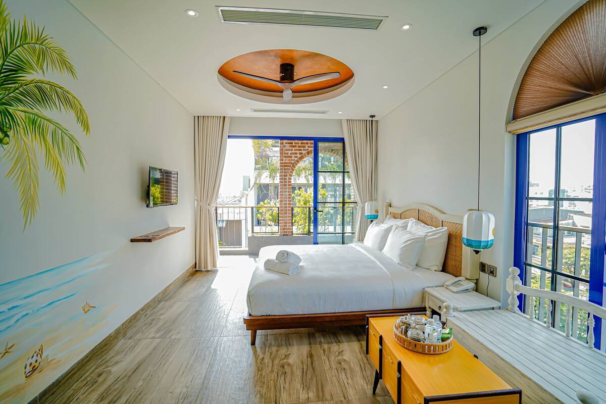 Habitación de hotel con una cama grande, cabecera de madera, una mesita con una bandeja de objetos, ventilador de techo y vistas al exterior a través de amplios ventanales.