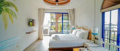 A hotel room with a large bed, a wooden headboard, a small table with a tray of items, a ceiling fan, and a view of the outside through large windows.