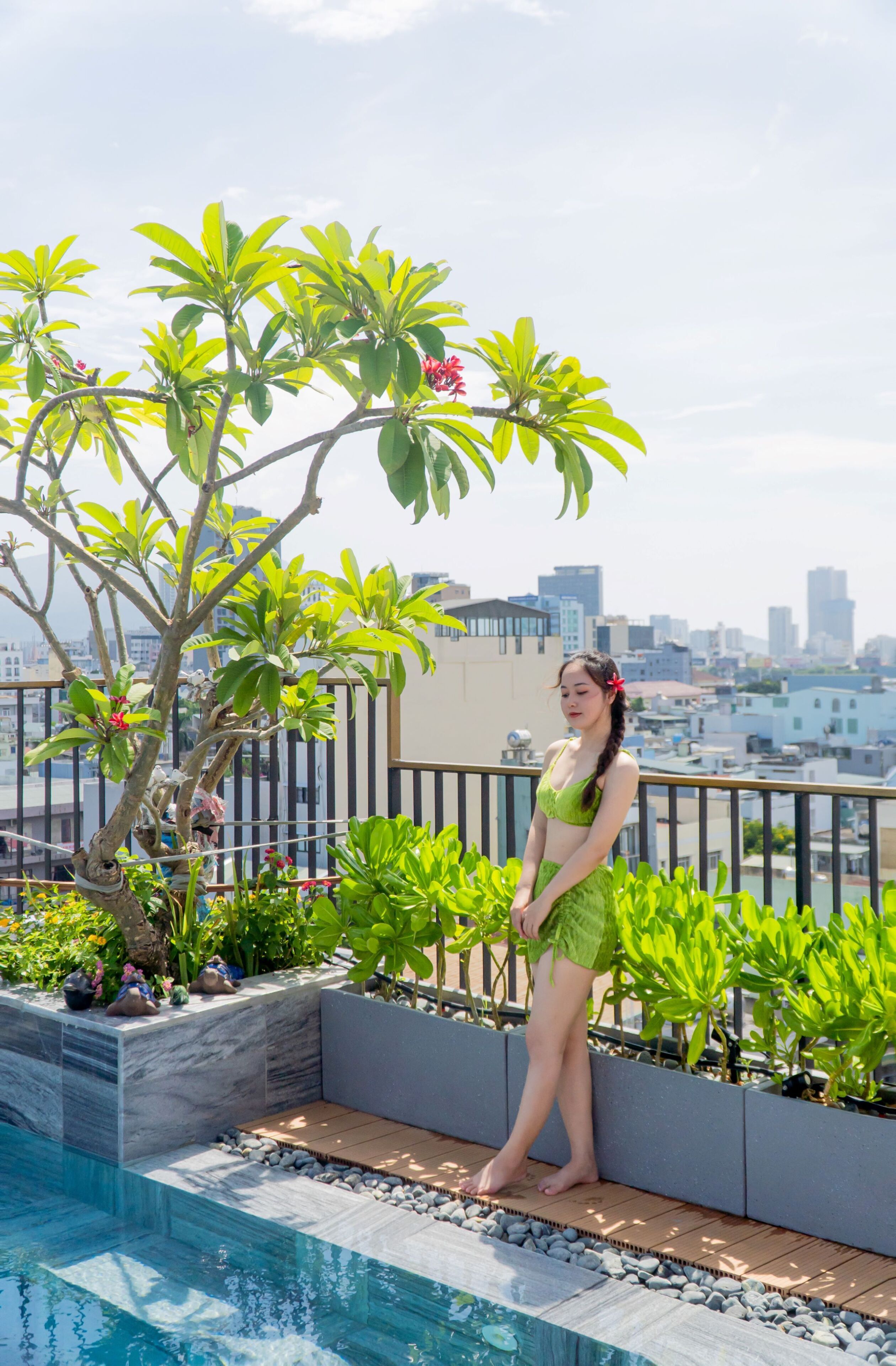 Mulher de fato de banho verde em frente a uma piscina, com paisagem urbana ao fundo.