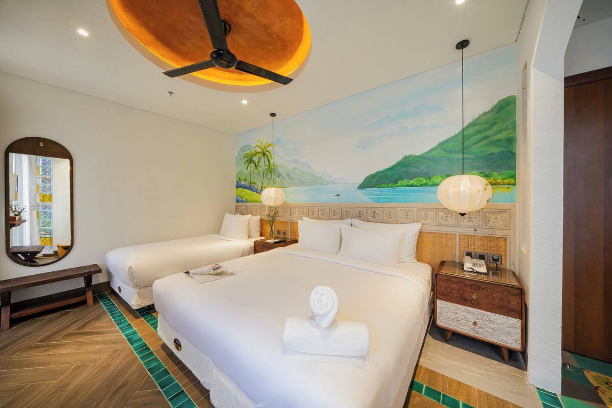 Habitación de hotel con dos camas, un mural grande de paisaje tropical, un ventilador de techo y una mesita de noche con lámpara.