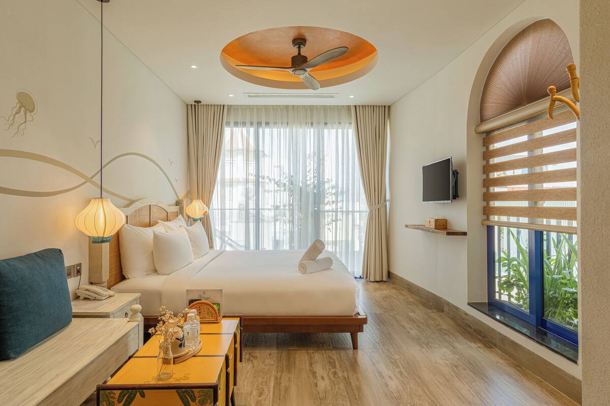 Habitación de hotel con una cama grande, un escritorio con lámpara, un televisor en la pared y una ventana con persianas.