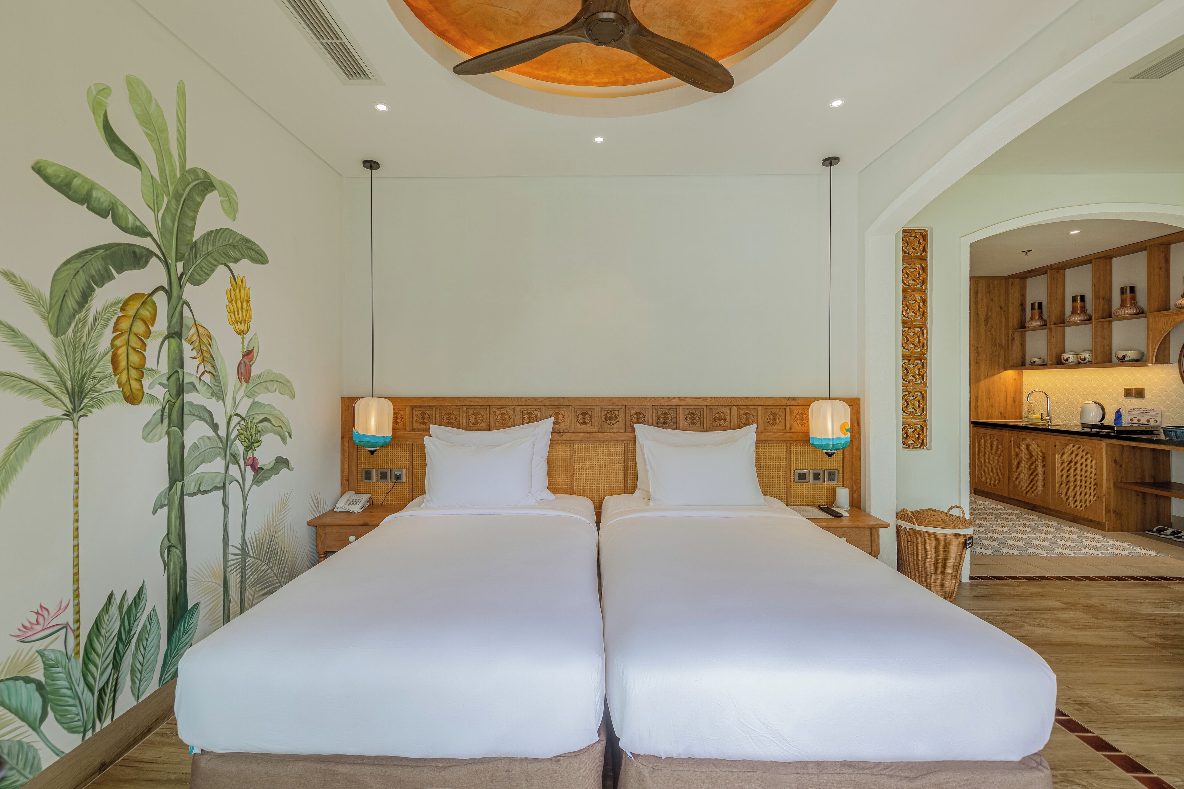 A hotel room with two beds, a ceiling fan, a kitchen area, and a mural of tropical plants.