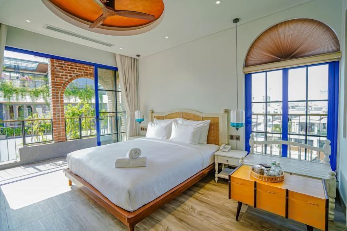 Habitación de hotel con una cama grande, un escritorio con lámpara y vistas a la ciudad a través de amplios ventanales.