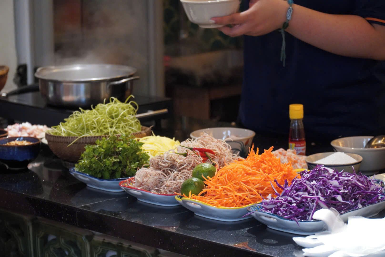On peut voir une variété de légumes frais et d’ingrédients sur un plan de travail de cuisine, notamment des carottes râpées, du chou et des herbes aromatiques.