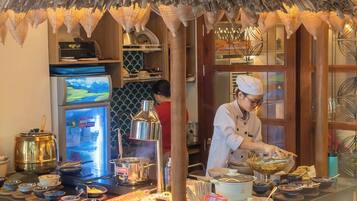 A restaurant with a thatched roof, a chef preparing food, and a variety of dishes displayed on the counter.