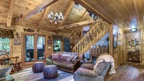 A wooden cabin interior with a sofa, armchair, and ottoman, a staircase, and various wall decorations.