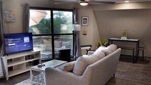 Una sala de estar con un sofá gris, un televisor sobre un mueble bajo, una mesita y una ventana con cortinas.