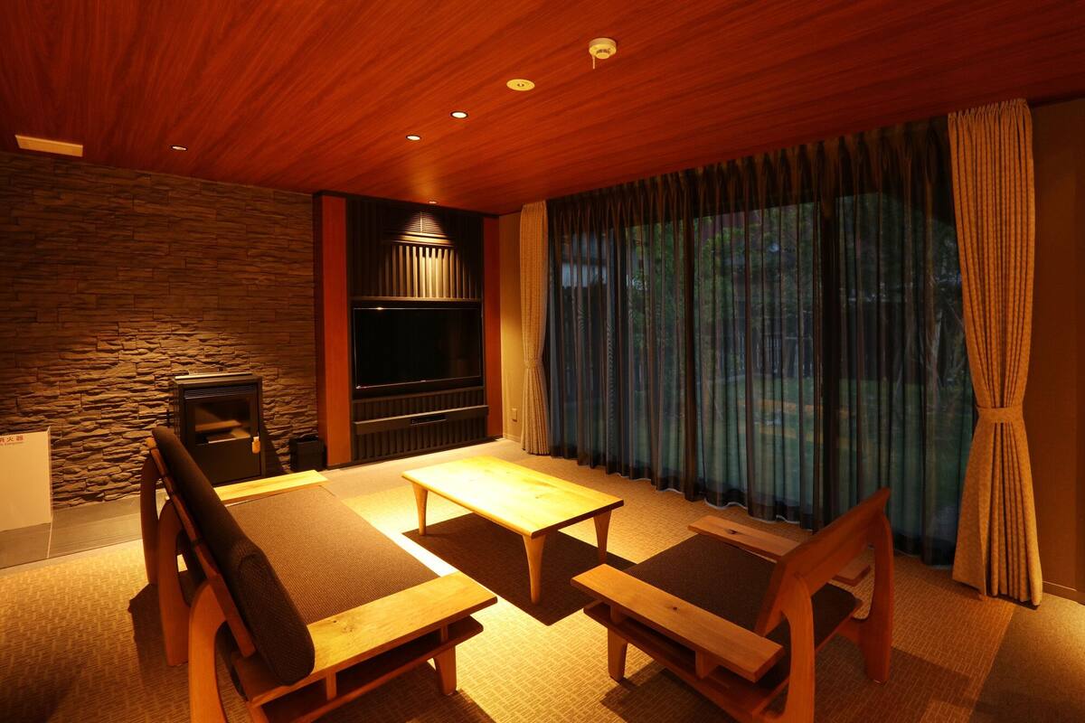 A cozy room with a stone wall, a fireplace, a large window with curtains, and a wooden dining table with chairs.