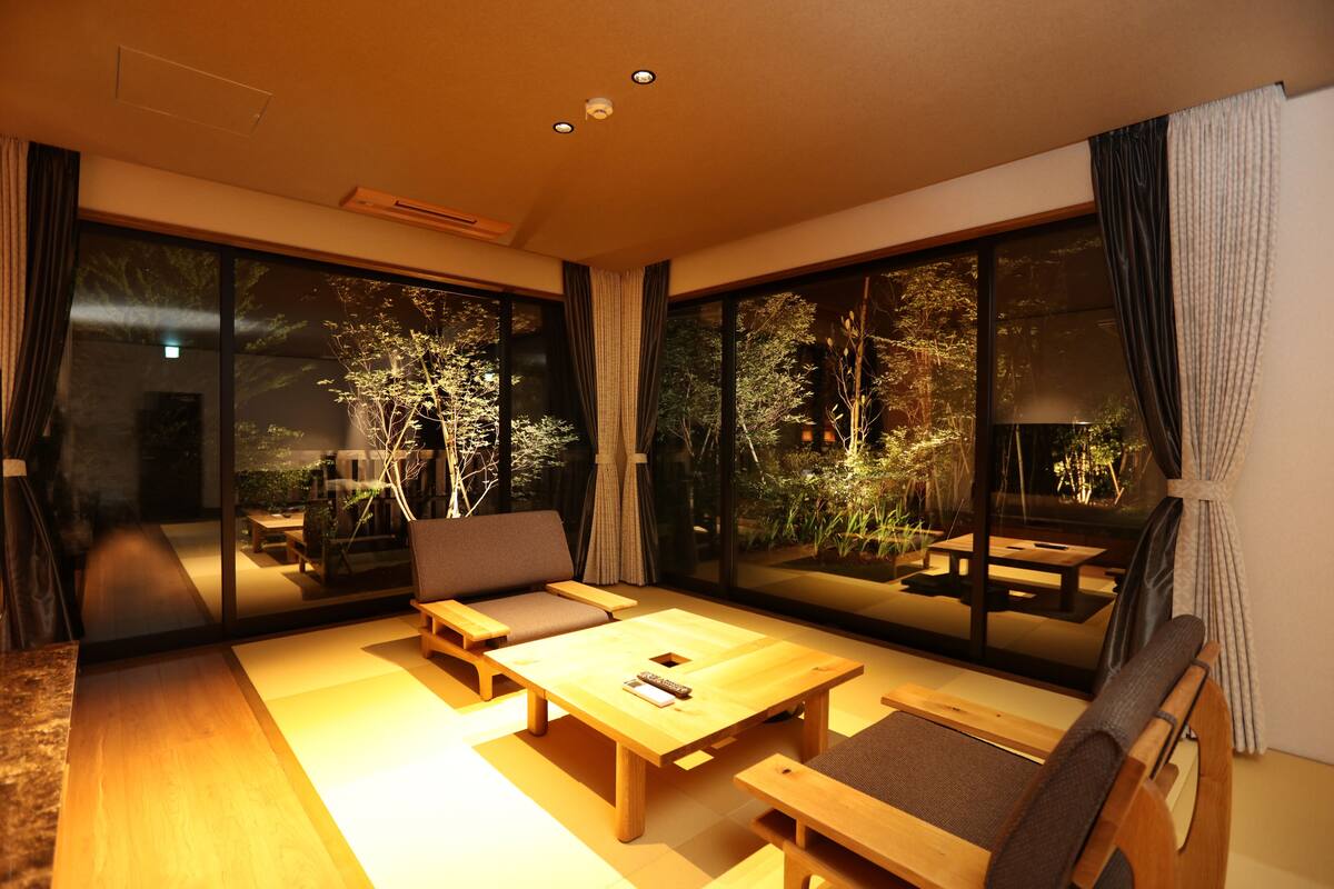 A modern living room with a wooden coffee table, sofa, and chairs, large windows with curtains, and a view of a garden.