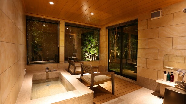 Un baño moderno con una bañera grande, asientos de madera y amplias ventanas con vistas a la vegetación.
