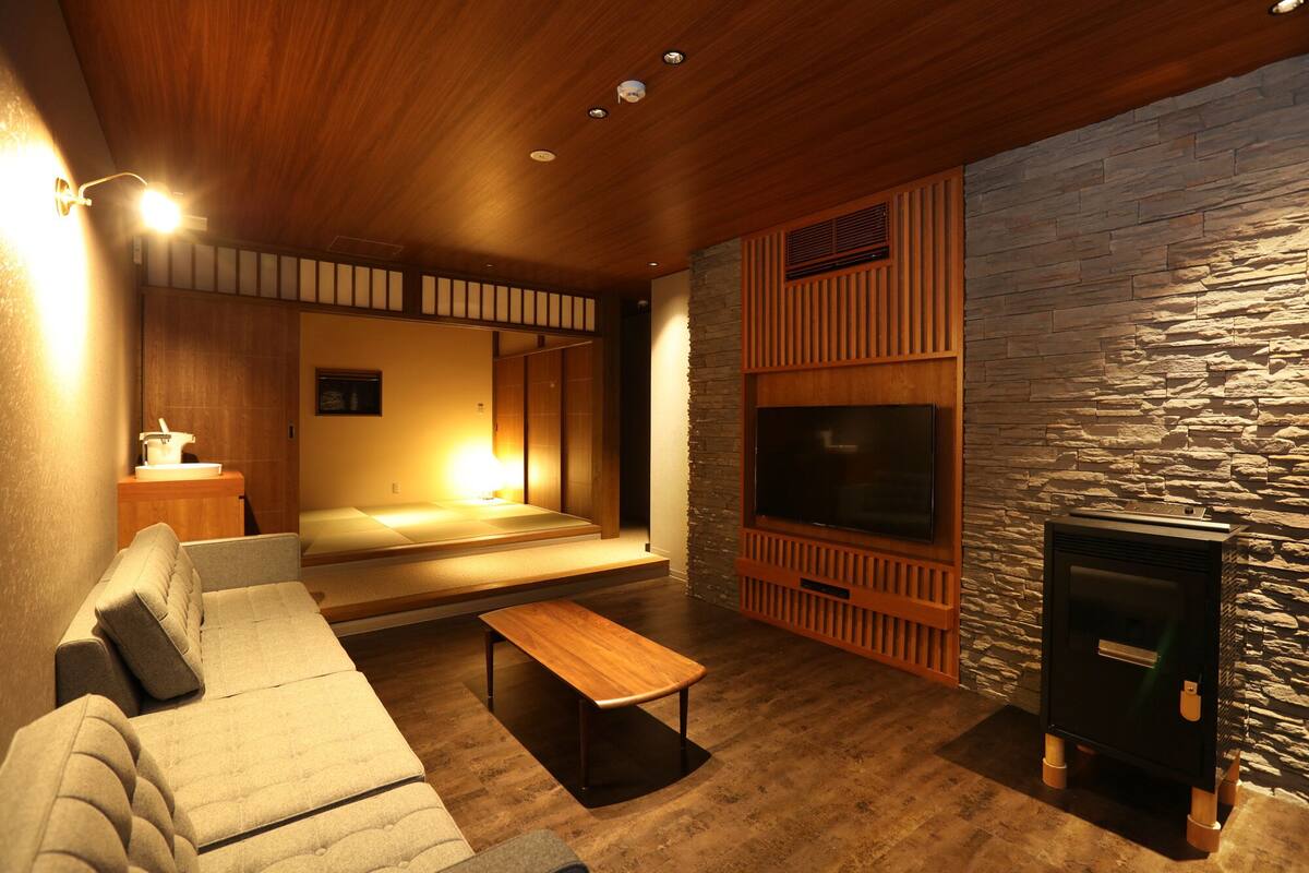 A cozy living room with a sofa, a wooden coffee table, a stone wall, a fireplace, and a television.