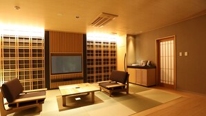 Una habitación tradicional japonesa con suelo de tatami, una mesa baja, dos sillas, un televisor y puertas correderas.