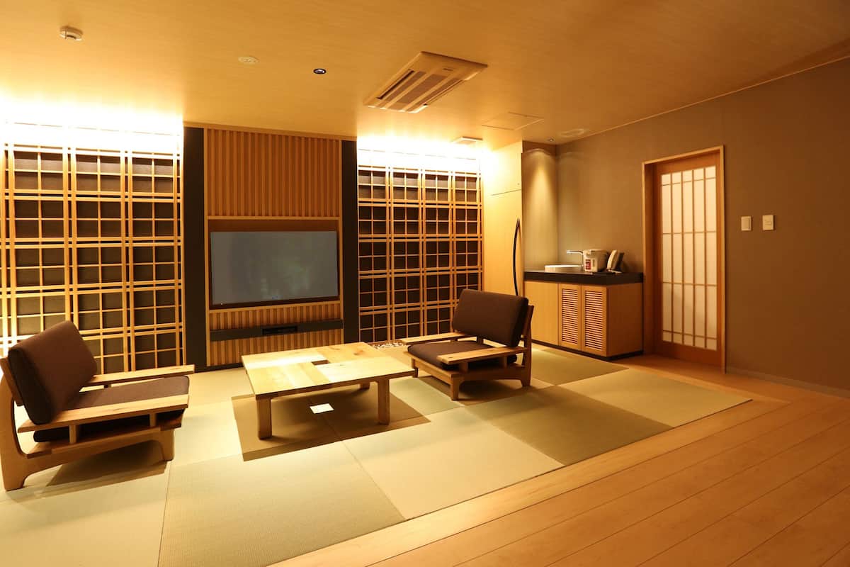 A traditional Japanese-style room with tatami flooring, a low table, two chairs, a television, and sliding doors.