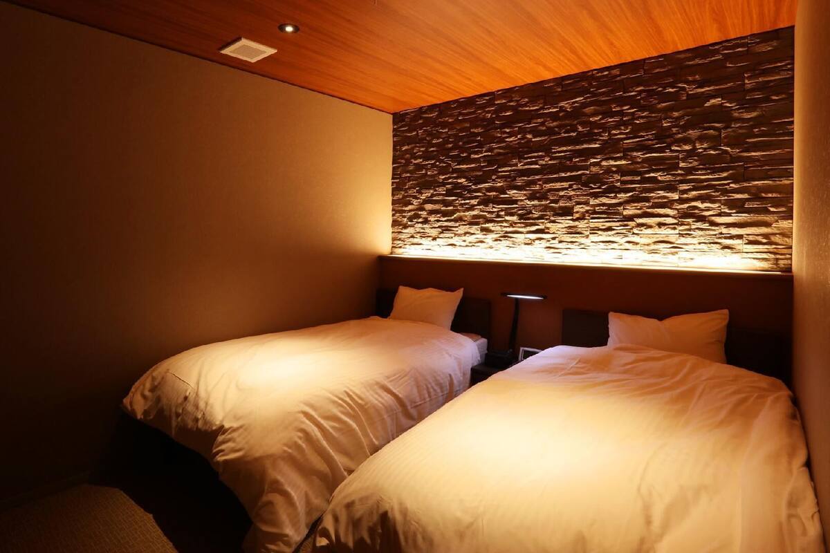 A hotel room with two single beds, a stone wall, and a ceiling light.