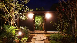 Un sendero conduce a una construcción de madera con un acceso cubierto, rodeada de vegetación e iluminada con luces.