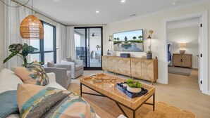 A modern living room with a sofa, coffee table, and a large TV. There is a dining area visible through a glass door.