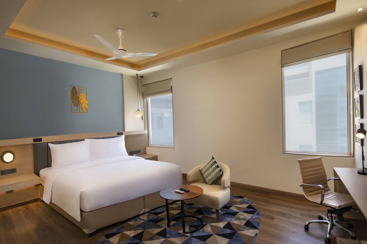 A hotel room with a bed, a desk, a chair, a small table, and a ceiling fan.